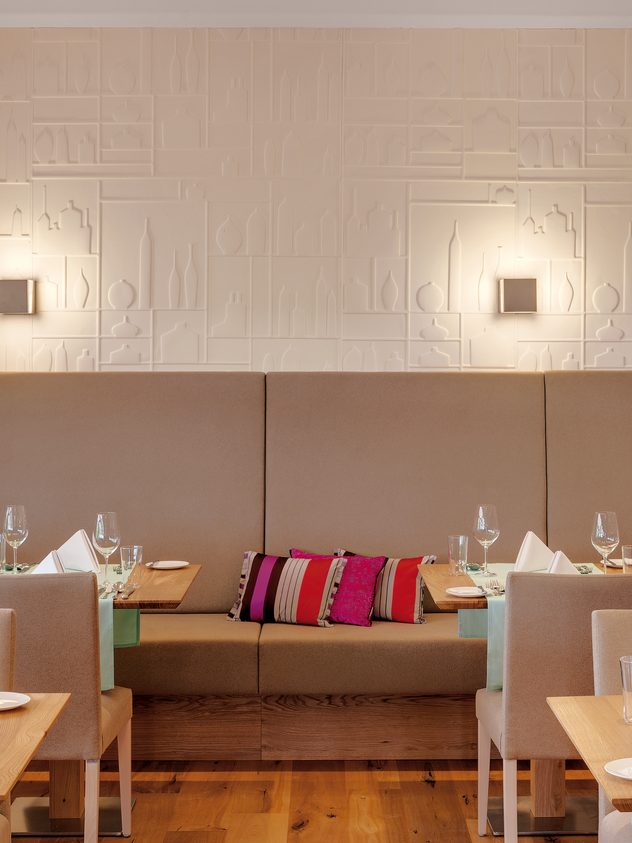 Restaurantbereich mit gedeckten Tischen, Stühlen und gepolsterter Sitzbank an einer beleuchteten Wand.
